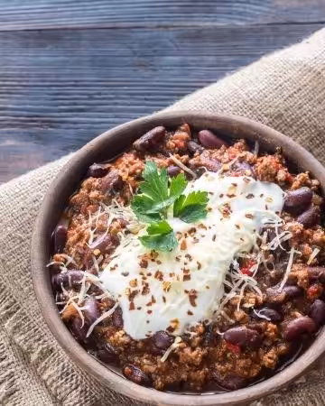 An earthenware bowl of chili con carne, topped with sour cream, grated cheese and parsley.