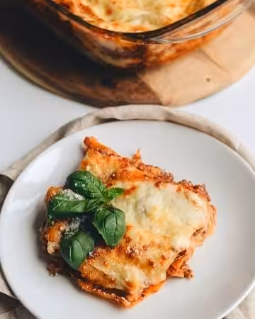 A slice of golden brown lasagna garnished with basil on a white plate with a glass baking dish full of lasagna in the background..
