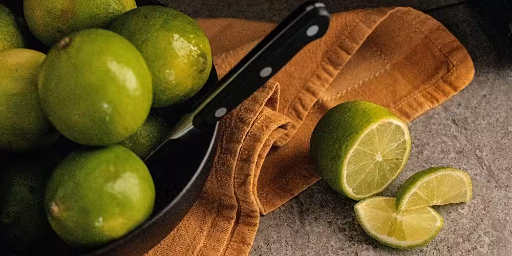A sliced lime next to a black plate full of whole limes.