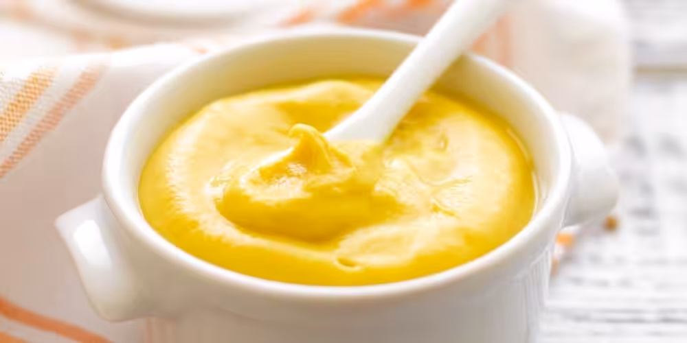 Closeup of a white ramekin full of yellow mustard with a small white spoon.