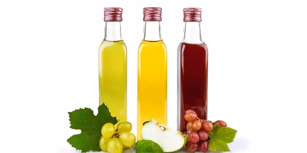 Closeup of 3 unlabelled glass bottles of vinegar with grapes and apple slices in front.