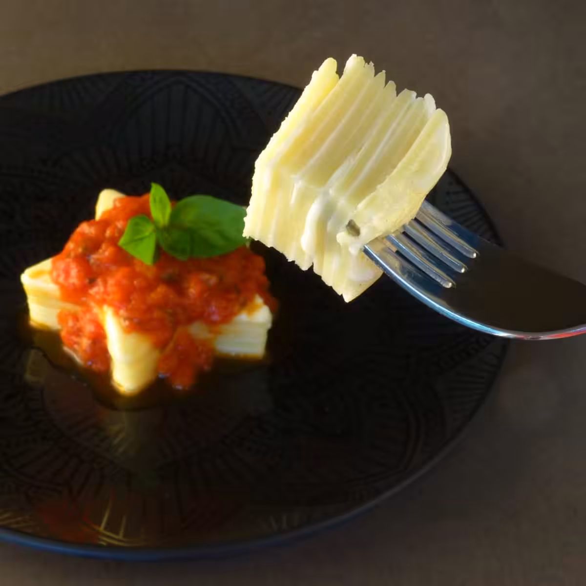 Closeup of a piece of Inside Out Lasagne on a fork with a star shaped tomato sauce Inside Out Lasagne on a dark plate in the background.